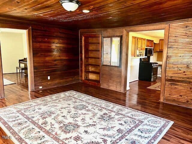 A room with polished wooden walls and ceiling, featuring a patterned rug on a wooden floor. There is a built-in bookshelf, a doorway leading to a kitchen with wooden cabinets and appliances, and a piano partially visible in an adjoining room.