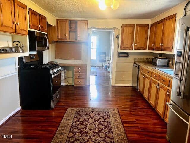 A spacious kitchen with wooden cabinets and polished hardwood floors. It features a gas stove, dishwasher, microwave, and a refrigerator. A rug is placed in the center, and a doorway at the back leads to another room.