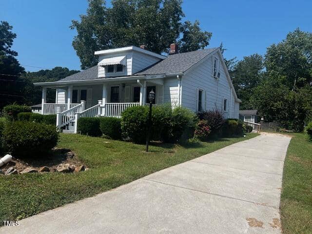 A white, single-story house with a gray roof, front porch, and black awnings. It's surrounded by shrubs and trees, with a concrete driveway leading up to it. The sky is clear and blue.