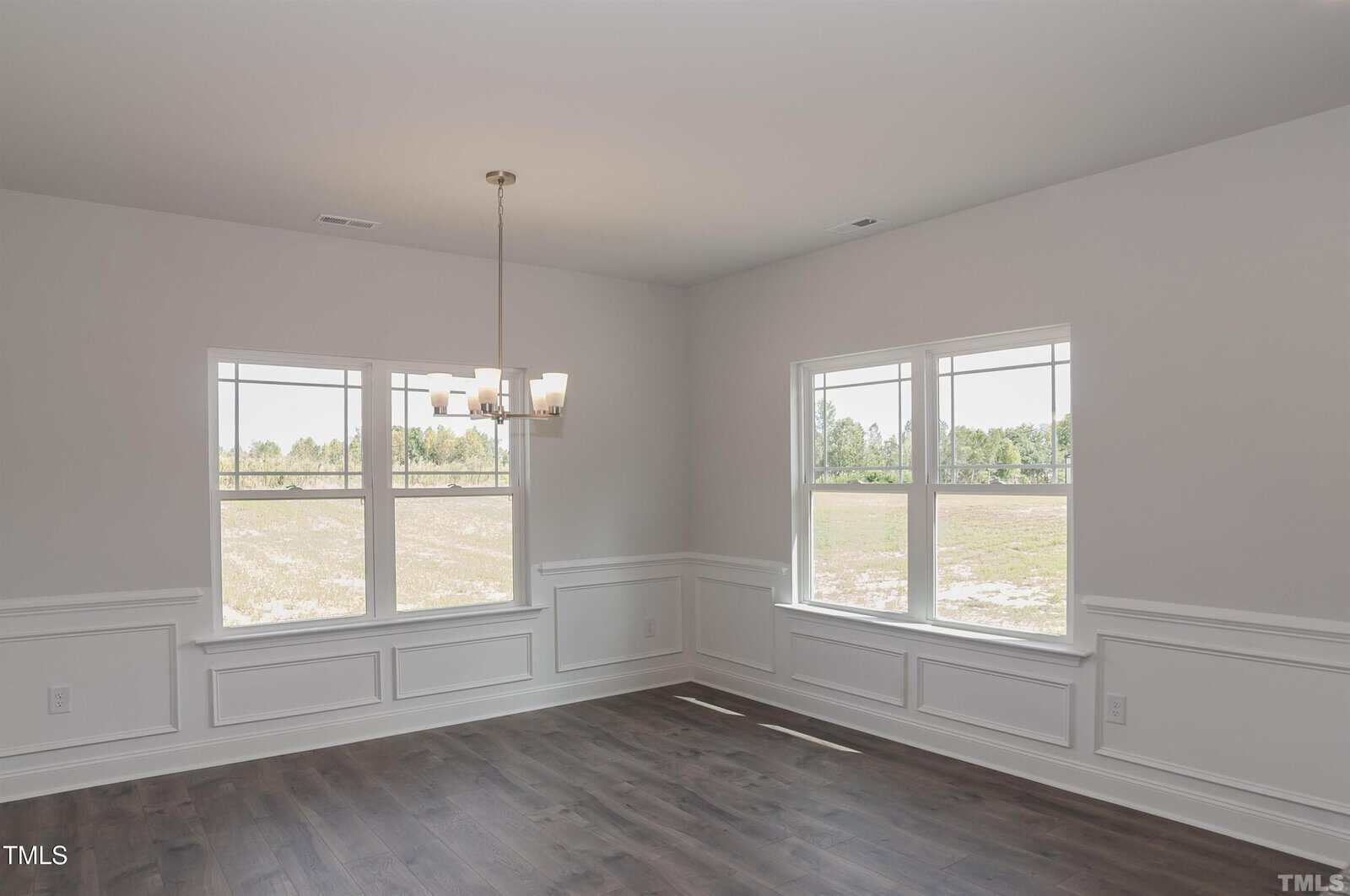 A bright, empty room with white walls, large windows, dark wood floors, and a modern chandelier hanging from the ceiling. Natural light streams in, highlighting the clean and spacious feel.
