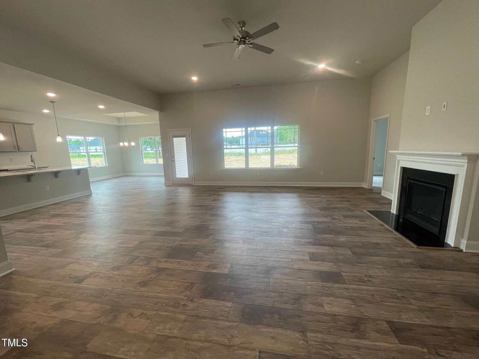 A spacious, empty living area with wood-look flooring, neutral-colored walls, large windows letting in natural light, a ceiling fan, a modern fireplace, and an open view of the adjacent kitchen and dining area.
