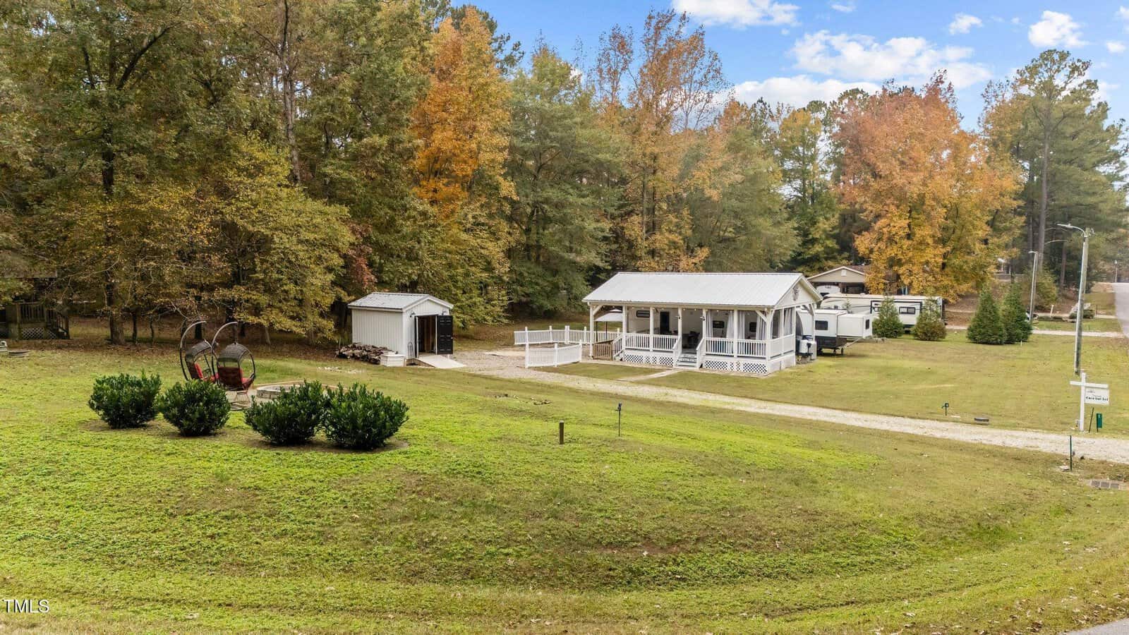 A small white house with a front porch sits on a green lawn surrounded by trees with autumn foliage. There is a gravel driveway, a small shed, and a swing chair on the grass near trimmed bushes.