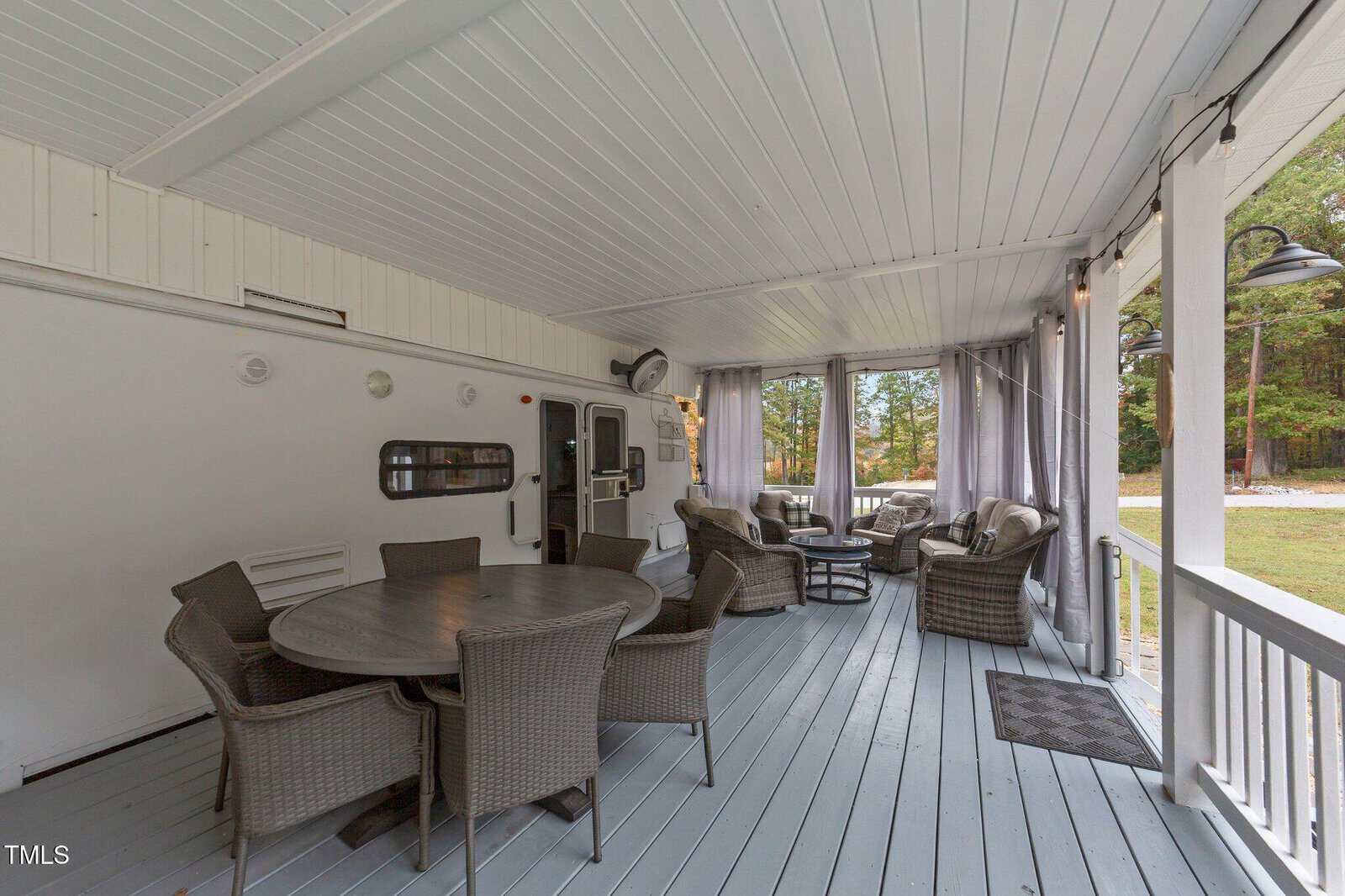 Covered outdoor patio with gray decking, round dining tables with wicker chairs, lounge chairs, outdoor curtains, and a view of a grassy yard with trees in the background.