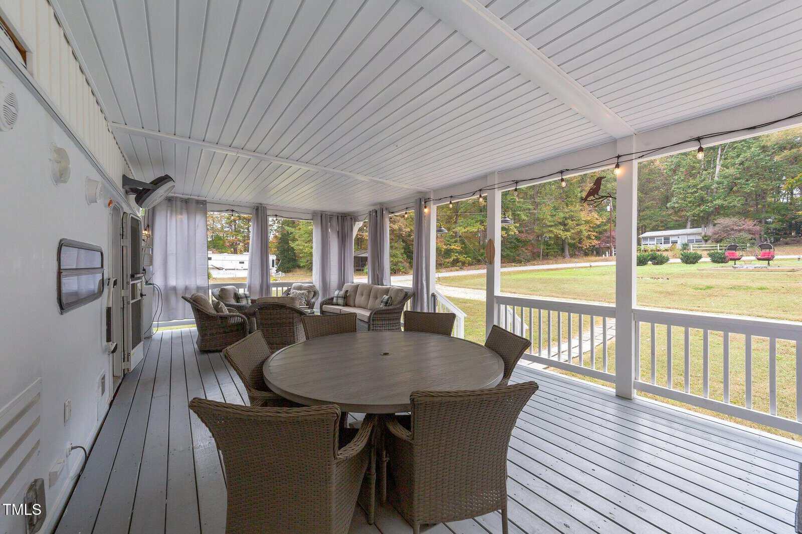 A covered porch with light gray wooden flooring and white railings features a round table surrounded by six wicker chairs. The porch overlooks a grassy yard with trees in the background.
