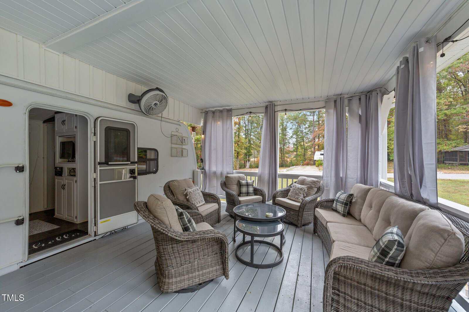 Screened-in porch with wicker furniture, gray cushions, plaid pillows, a round glass coffee table, a ceiling fan, curtains, and a view of trees and lawn outside. Part of an RV or trailer is visible on the left.