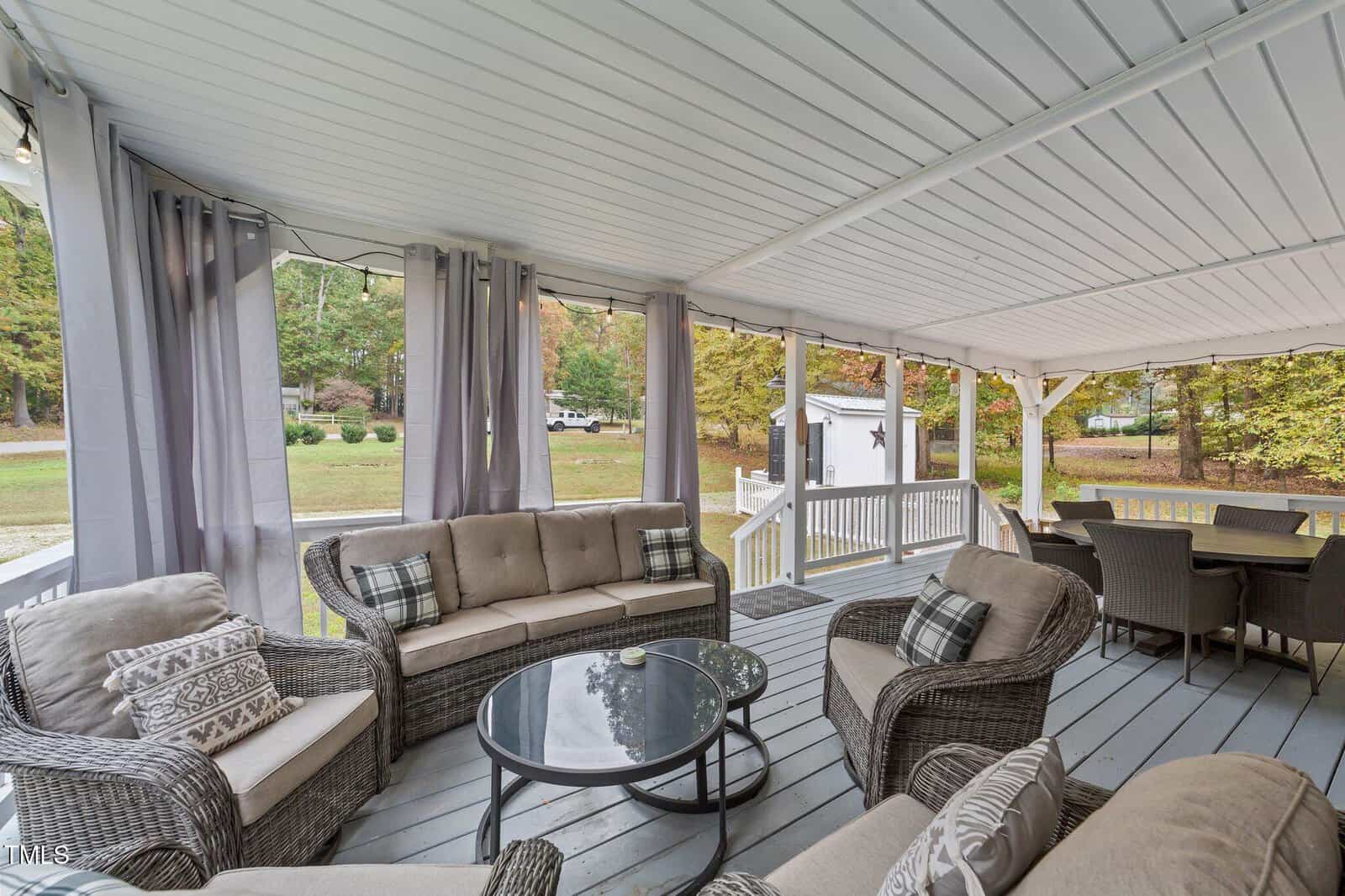 Covered patio with wicker furniture, gray cushions, and a round glass table. Dining set in the background. Gray curtains hang around the patio, overlooking a yard with trees and a shed.