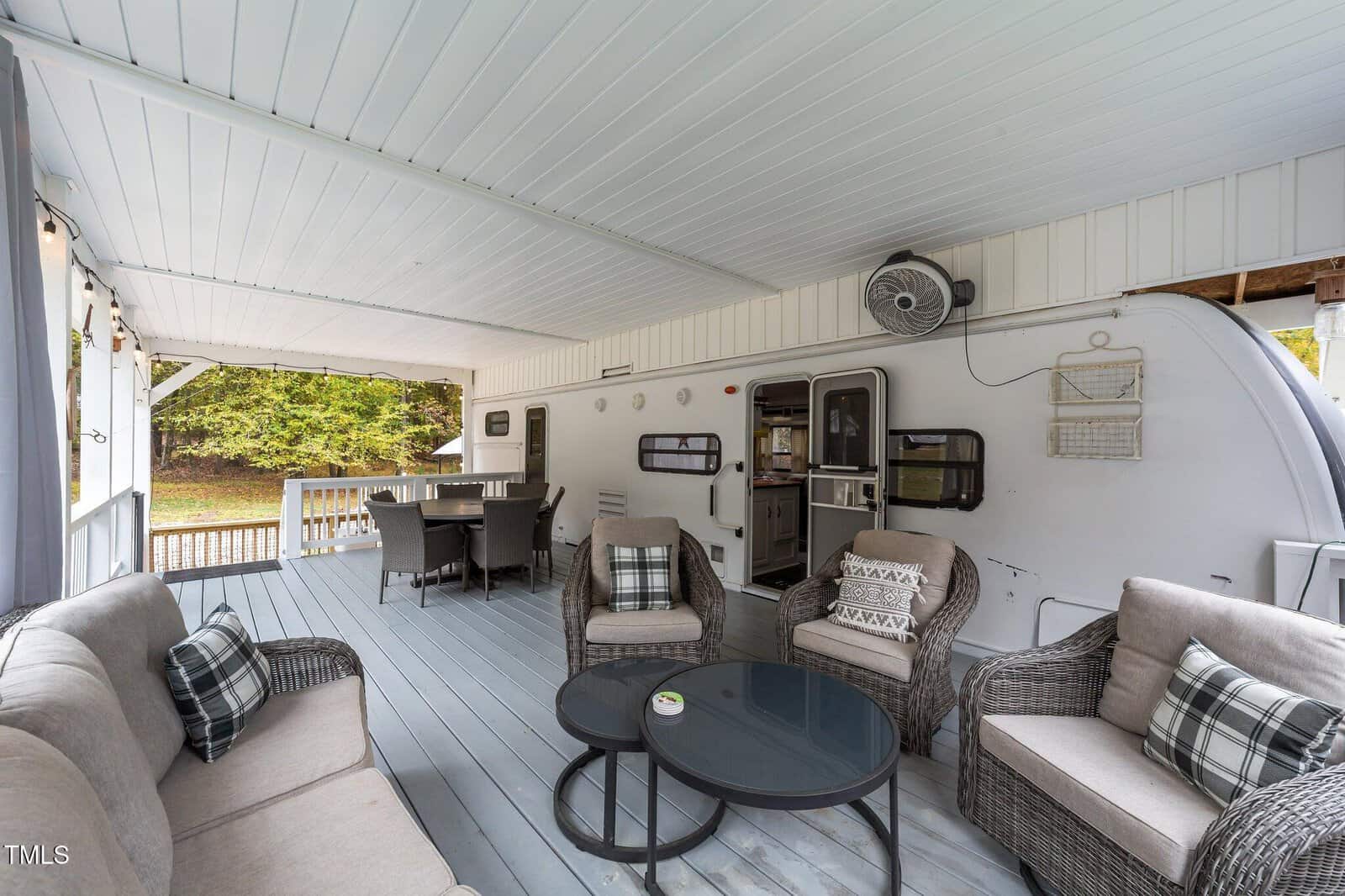 Covered porch with gray flooring, outdoor seating, and a coffee table. An RV is incorporated into the wall. There are potted plants, a ceiling fan, and a dining table in the background overlooking a green yard.