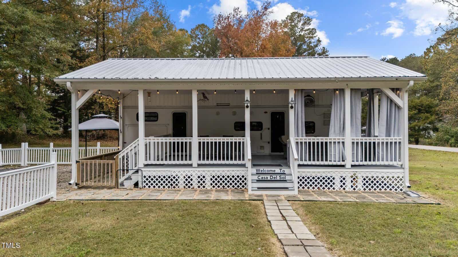 A white single-story house with a metal roof and a covered front porch sits on a grassy lawn. Trees with autumn foliage are in the background. Steps lead up to the porch, which has railings and outdoor lights.
