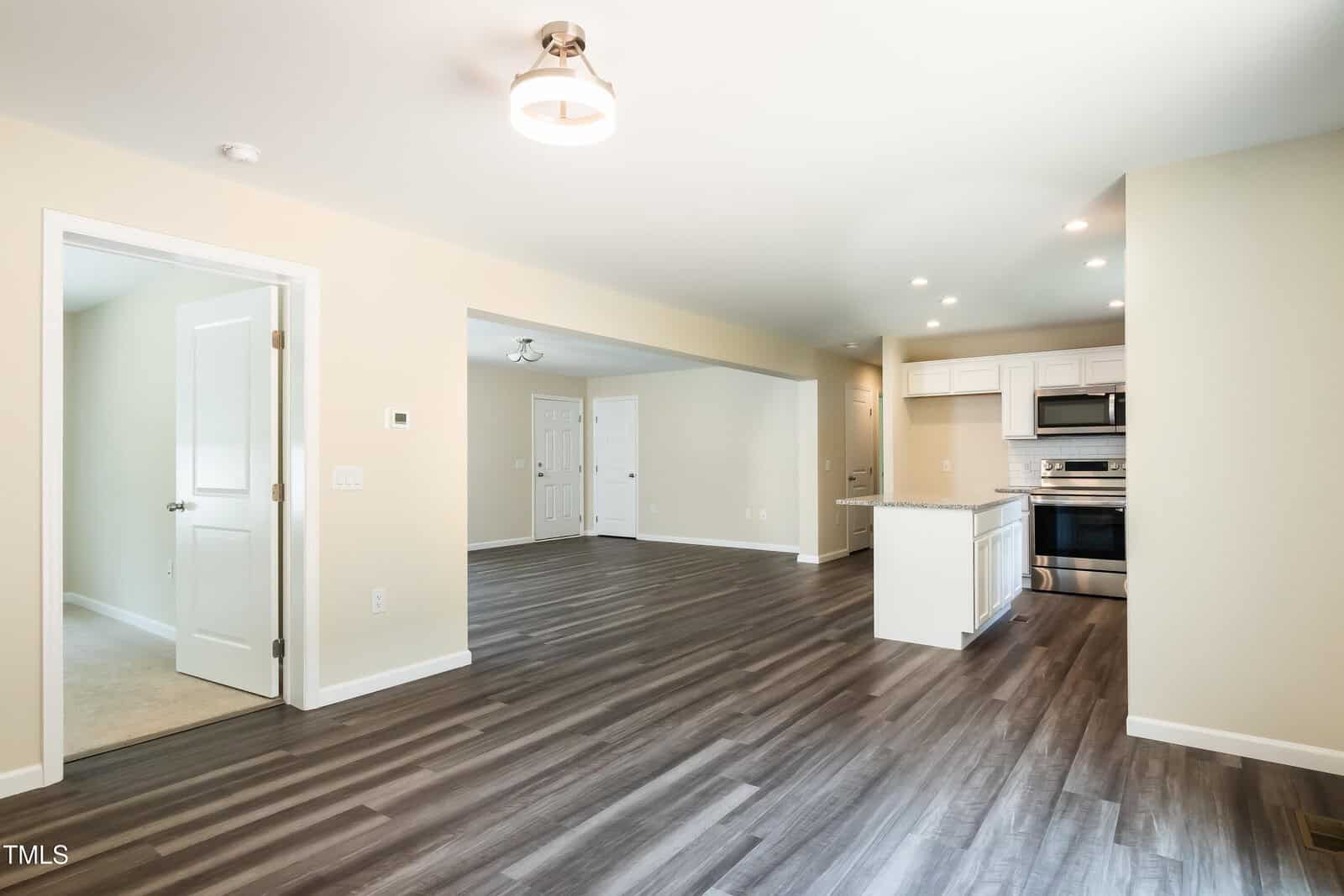 Open-concept living space with wood-style floors, beige walls, modern ceiling light, and a view of a kitchen with white cabinets and stainless steel appliances. A door leads to a carpeted room on the left.