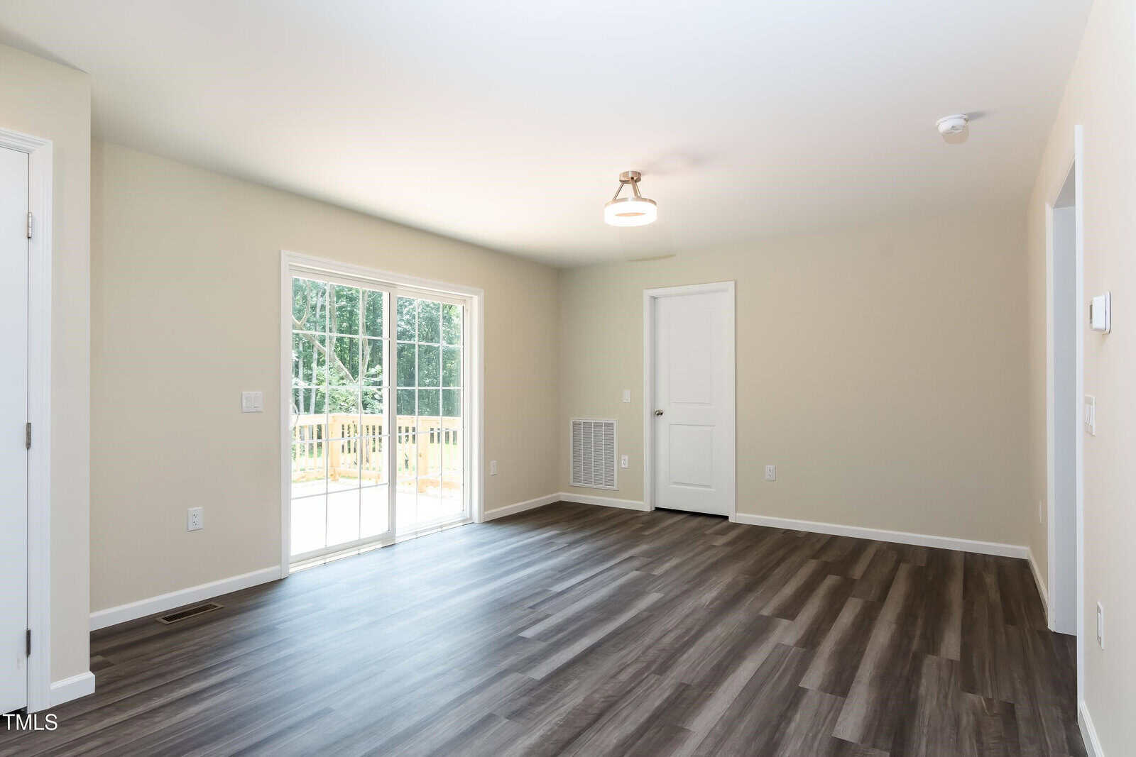 Bright empty room with light beige walls, wood-style flooring, sliding glass doors leading outside, a white door, ceiling light, and a vent on the wall. Natural sunlight enters from the sliding doors.