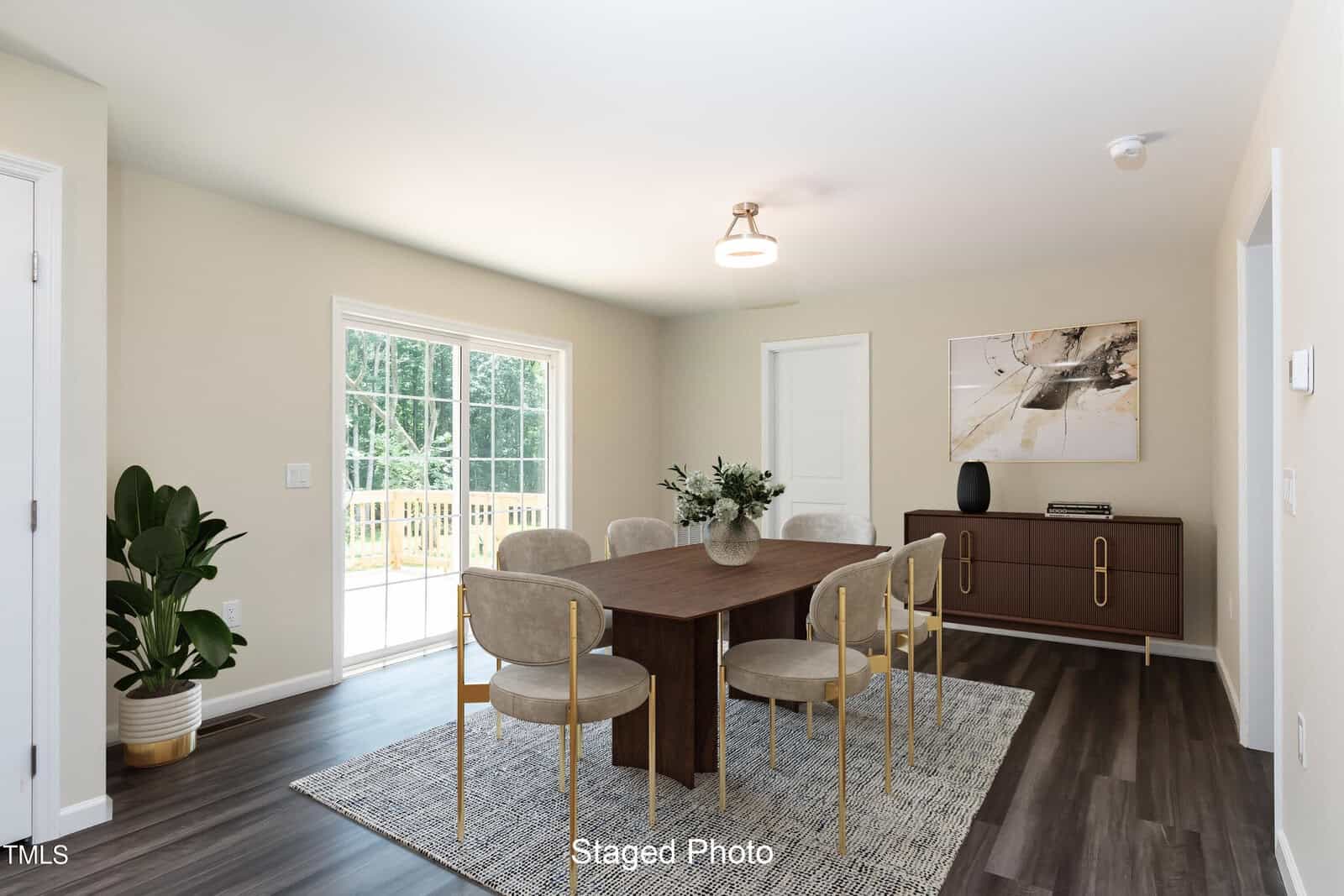 A modern dining room with a wooden table, six chairs, a sideboard, potted plant, abstract wall art, and large glass doors leading outside. The floor is dark wood, and a light rug is under the table.
