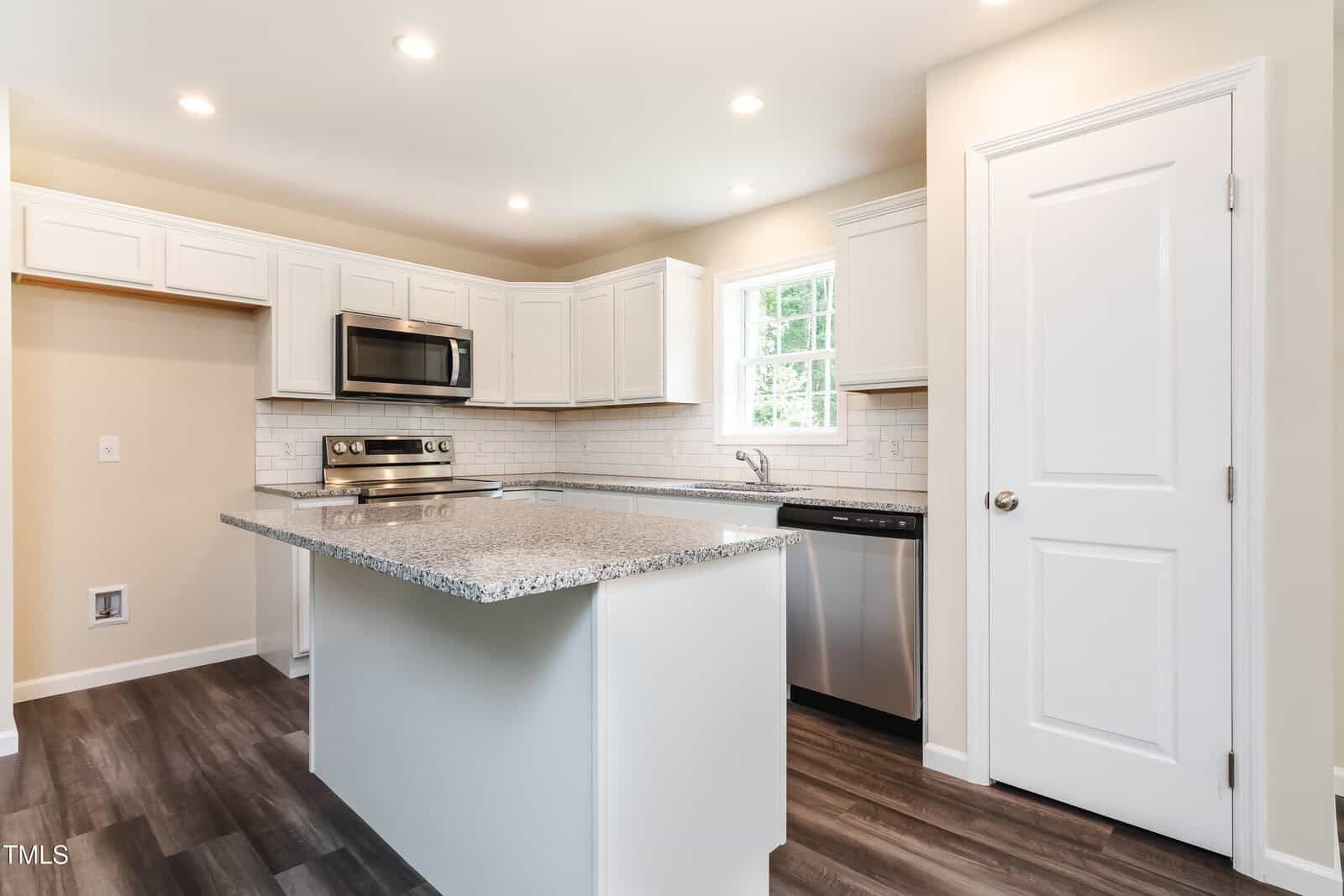 Modern kitchen with white cabinets, stainless steel appliances, gray granite countertops, a large center island, wood-look flooring, subway tile backsplash, and a window above the sink letting in natural light.