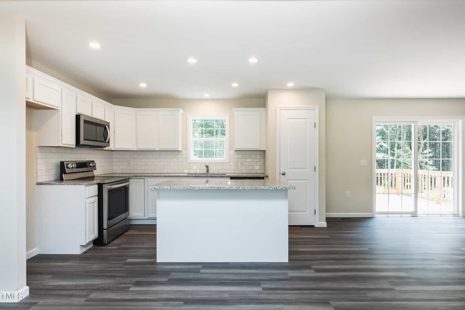 Modern, white kitchen with stainless steel appliances, granite countertops, a central island, subway tile backsplash, and wood-style flooring. Sliding glass doors open to a deck with a view of trees outside.