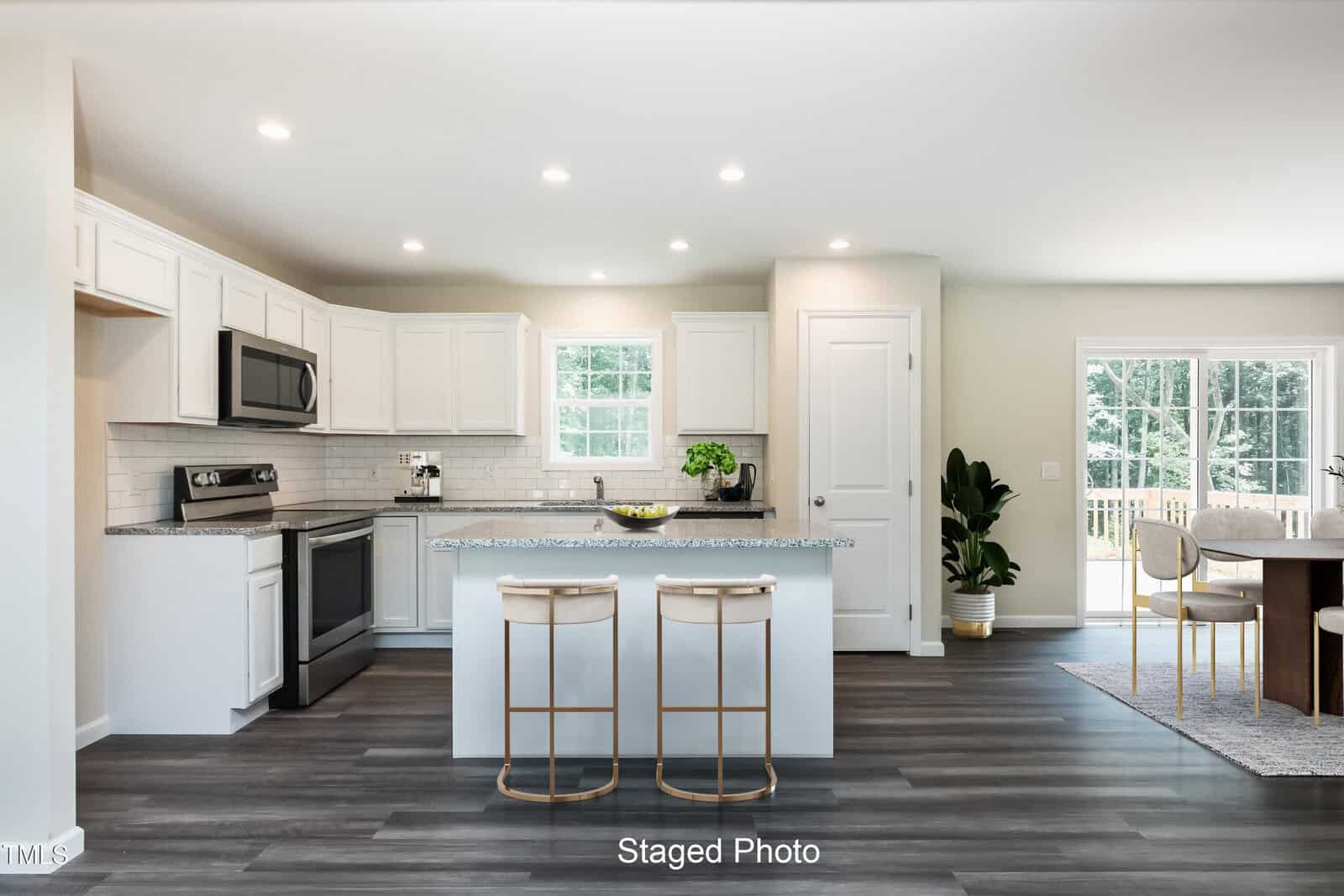 Modern kitchen with white cabinets, stainless steel appliances, an island with two stools, and a dining area. Large windows bring in natural light; a potted plant stands near the door. "Staged Photo" text at the bottom.