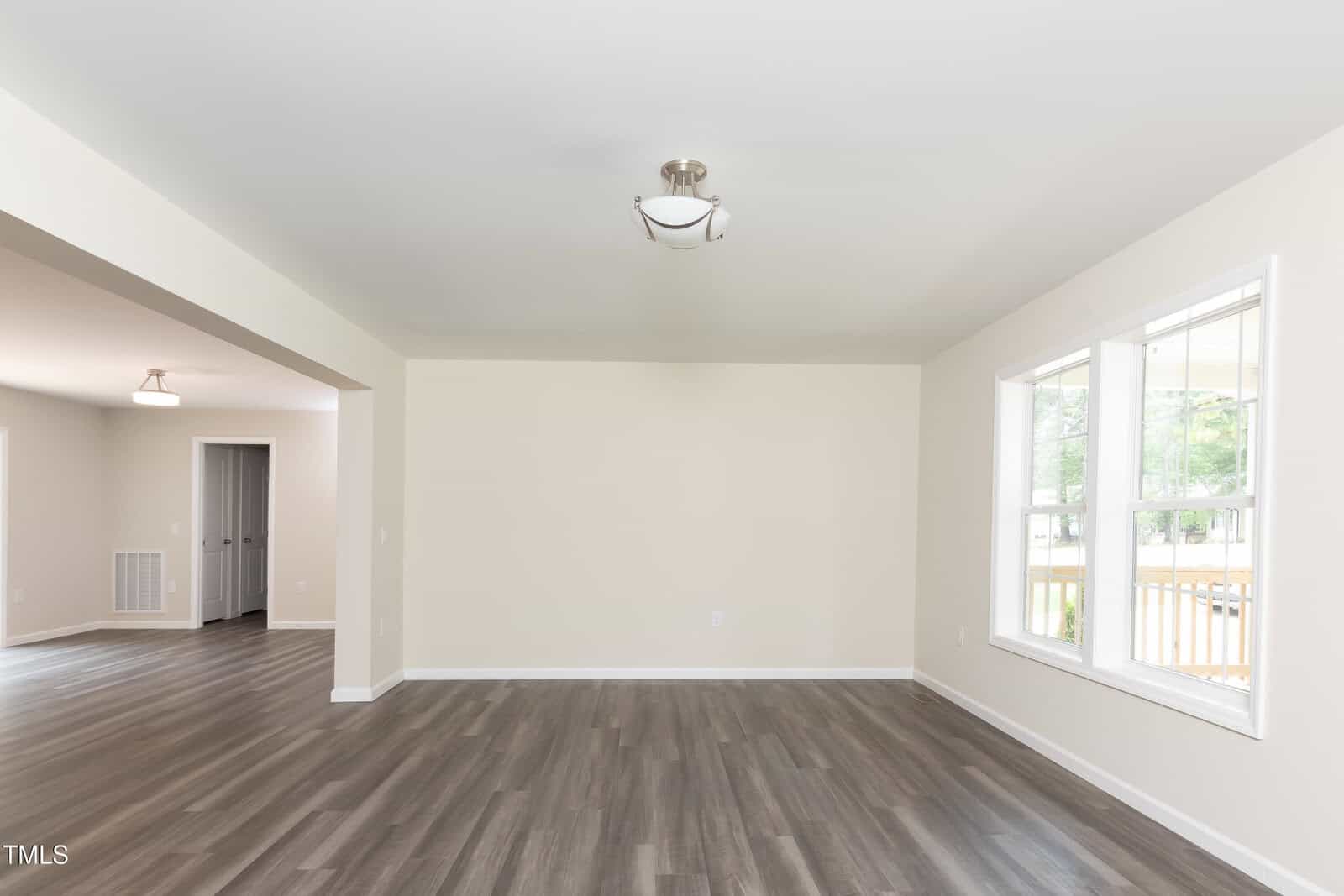 A bright, empty living room with light beige walls, large front window, modern ceiling light, and dark wood flooring. An open entryway leads to an adjacent room with similar flooring and lighting.