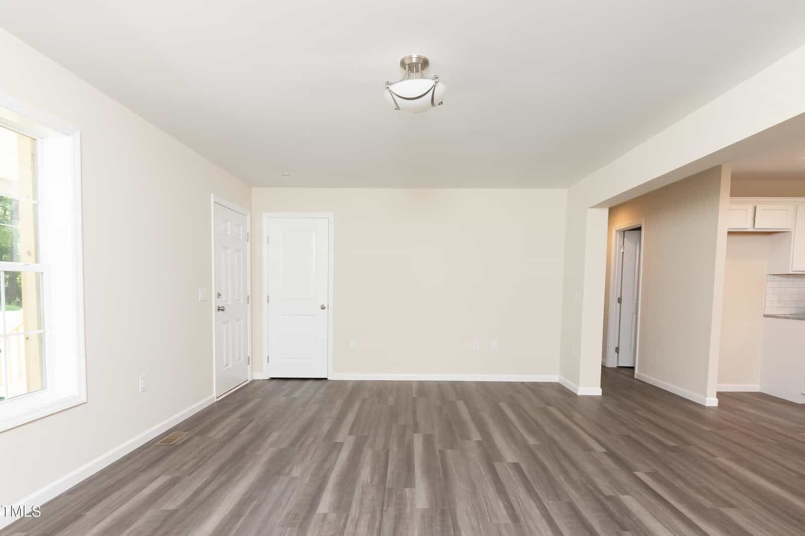 A bright, empty living room with light beige walls, wood-style flooring, a window on the left, two white doors at the back, and an open entryway to a kitchen area on the right.