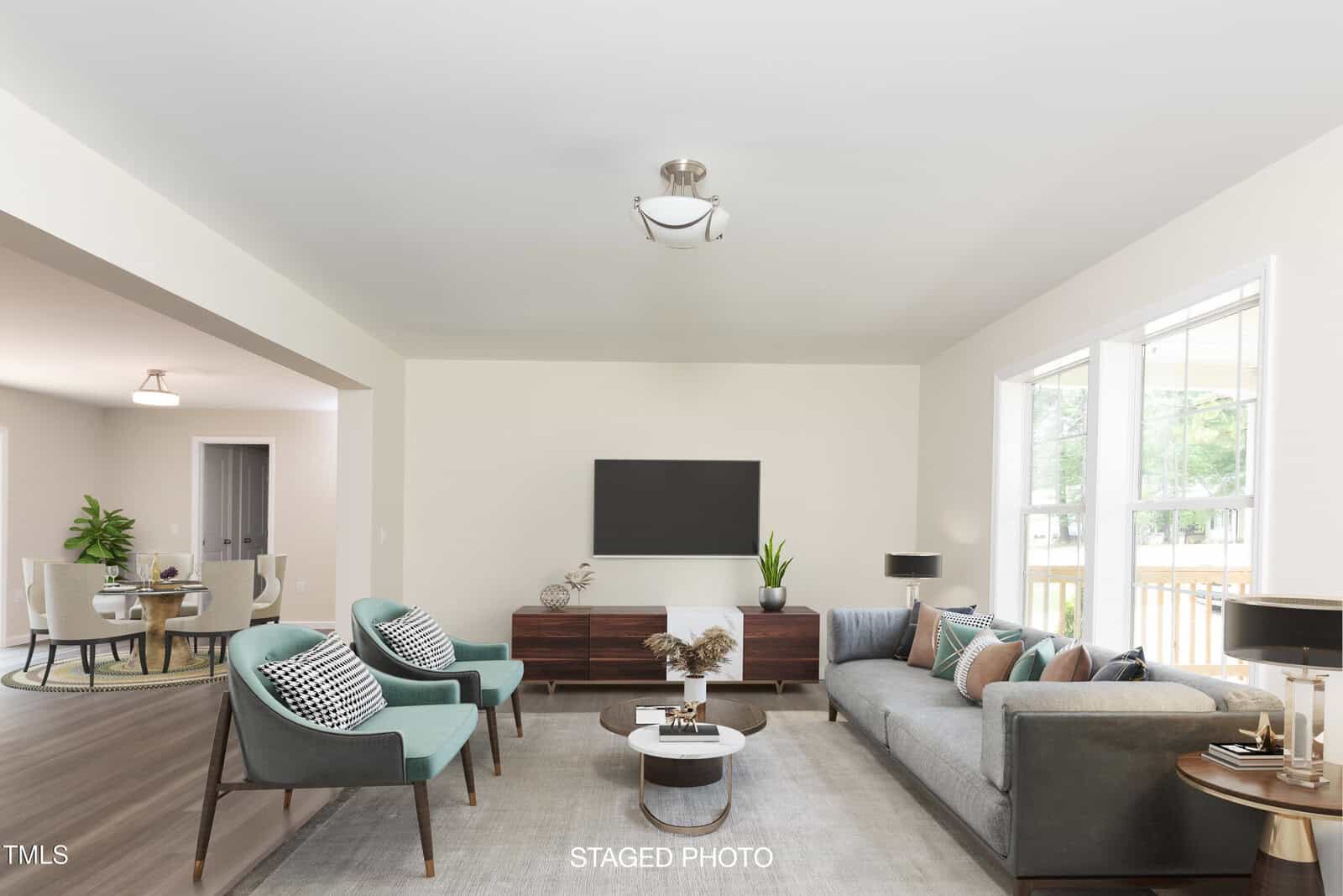 A modern, staged living room with a gray sofa, two teal armchairs, a wooden coffee table, a TV on a console, large windows, and a dining area visible in the background. Light walls and wood floors create a bright, open space.