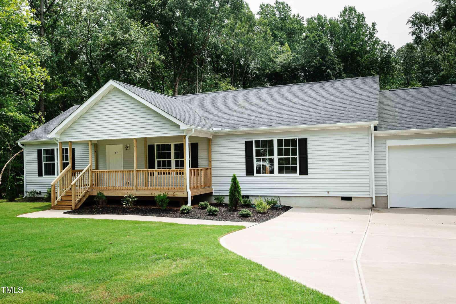 Single-story white house with black shutters, a covered front porch with wooden railings, attached garage, manicured lawn, small shrubs in front, and surrounded by tall trees in the background.