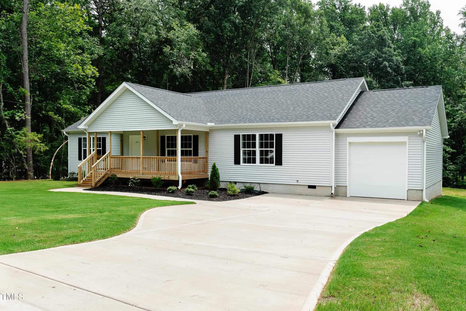 Single-story white house with a gray roof, front porch with wooden railings, attached garage, and a curved concrete driveway. The house is surrounded by a green lawn and trees in the background.