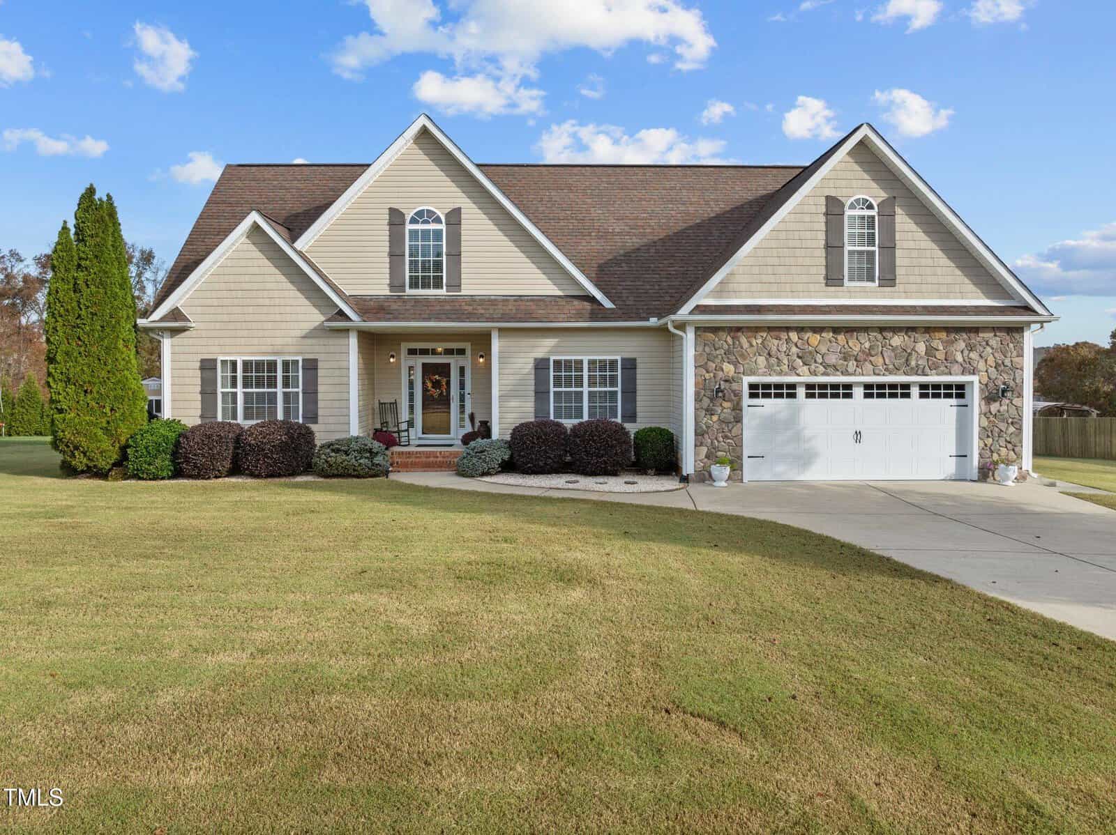 Located at 3519 Bluebonnet Drive in Wake Forest, this single-story house boasts a stone and beige siding exterior with a steep gable roof and a two-car garage. The front yard is adorned with neatly trimmed hedges and a manicured lawn, all under a clear blue sky with scattered clouds.