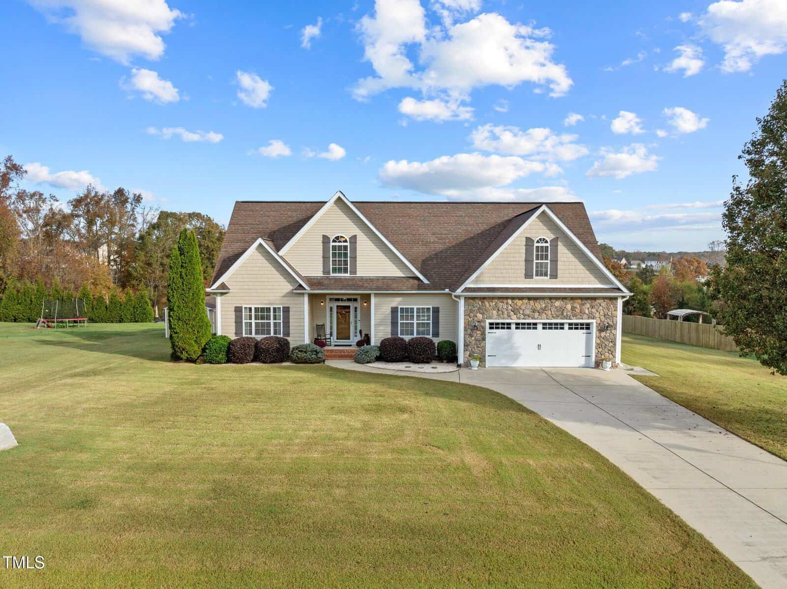 A suburban house with a gabled roof and stone accents sits on a large, well-manicured lawn. There's a double garage, a long driveway, and trimmed shrubs. The sky is partly cloudy, and trees are visible in the background.