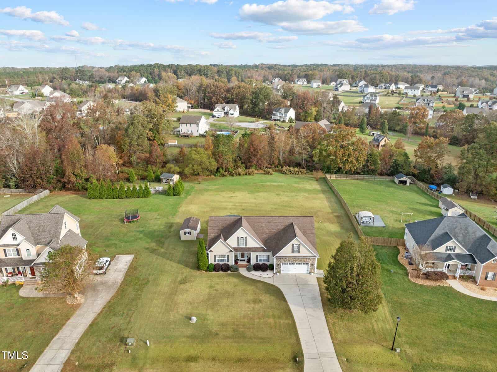Aerial view of a suburban neighborhood featuring a large house with a long driveway, surrounded by grass lawns and trees. Other houses are visible in the distance, with a scenic backdrop of autumn foliage and a partly cloudy sky.