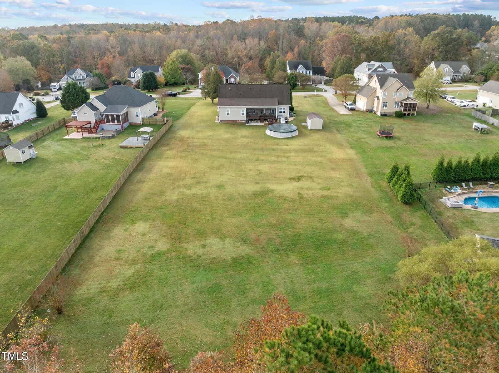 Aerial view of a suburban neighborhood with several large houses. Each house has a spacious backyard, some with pools or decks. The area is surrounded by trees under a partly cloudy sky.