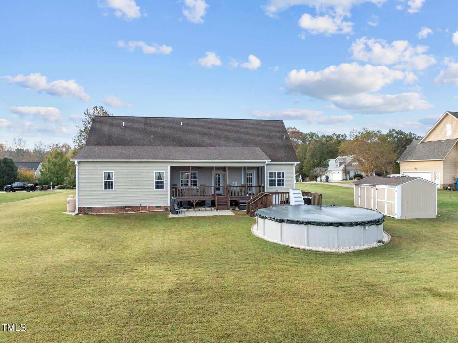 A suburban backyard with a large lawn featuring a light-colored house with a porch, an above-ground swimming pool, and a storage shed. The sky is partly cloudy, and the area is surrounded by other homes and trees.