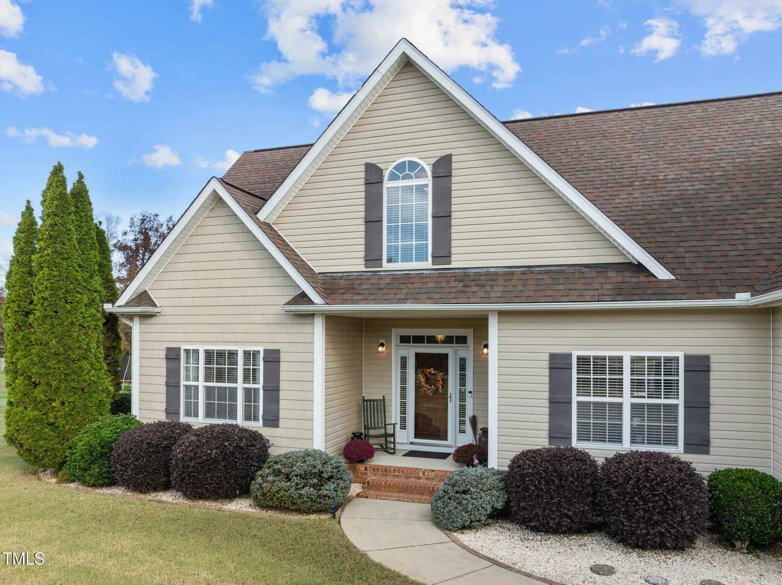 A beige, two-story house with a steep, pitched roof, dark shutters, and a front porch. The yard is landscaped with shrubs and a tall tree. The sky is blue with scattered clouds.