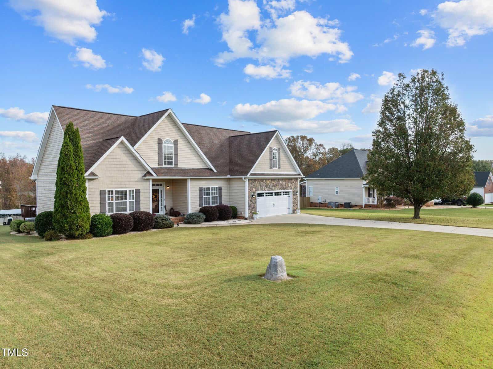 A beige house with a brown roof sits on a wide, manicured lawn under a blue sky with scattered clouds. The house features a large garage and neatly trimmed shrubs. A tall tree stands near the driveway, creating a picturesque suburban setting.