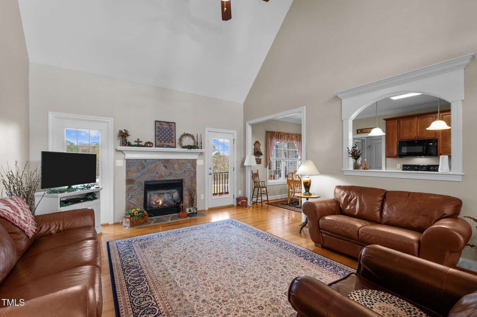 A cozy living room with high ceilings features a fireplace, a TV, and brown leather sofas. A patterned rug covers the wooden floor. A dining area and a kitchen with wooden cabinets are visible in the background. Plants and decor add warmth to the space.