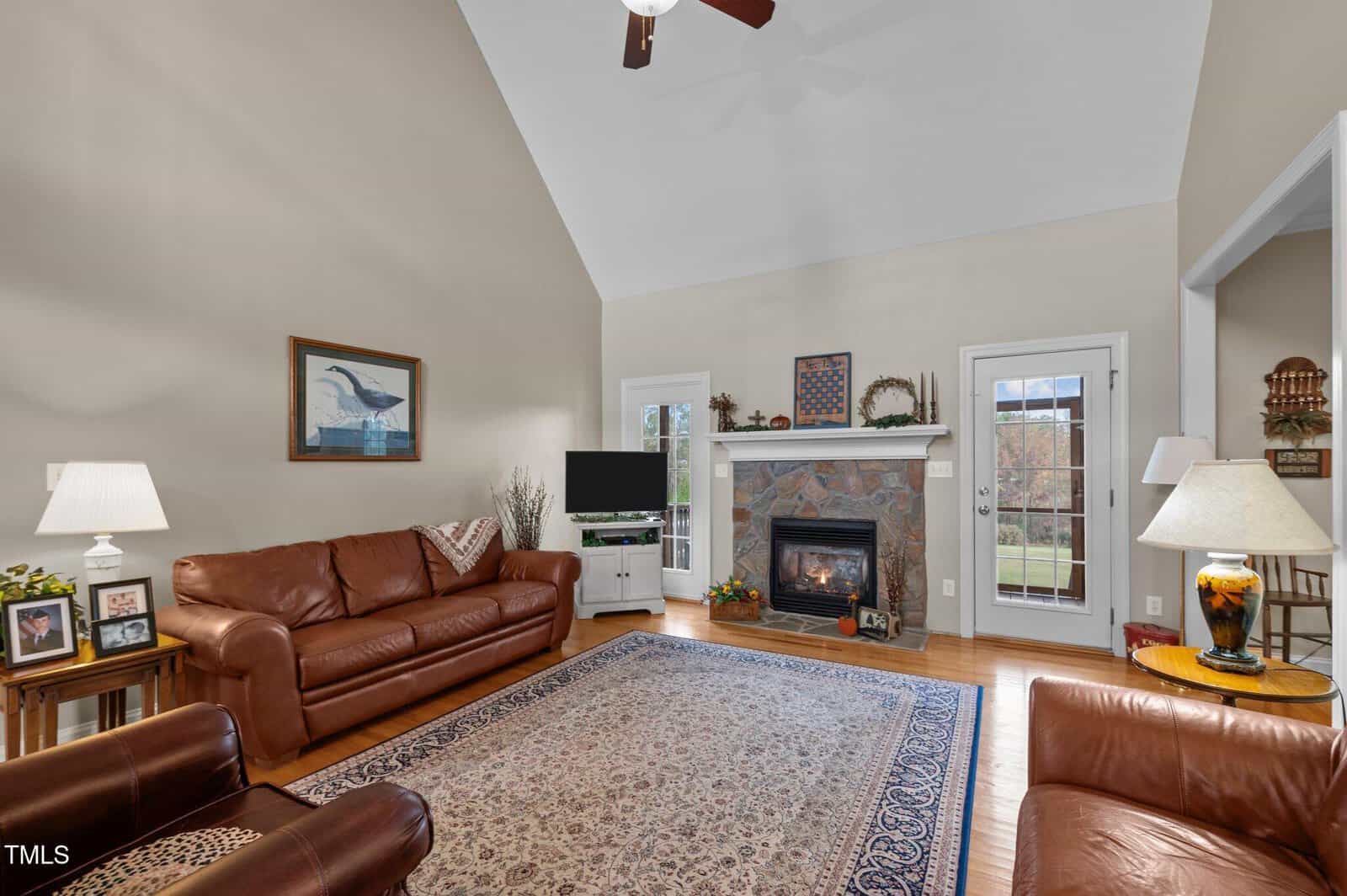 A cozy living room with high ceilings, leather sofas, and a patterned rug. A fireplace with decorative tiles is flanked by a door and a window. The room is warmly lit by lamps, and a ceiling fan hangs above. A painting and photos adorn the walls and tables.