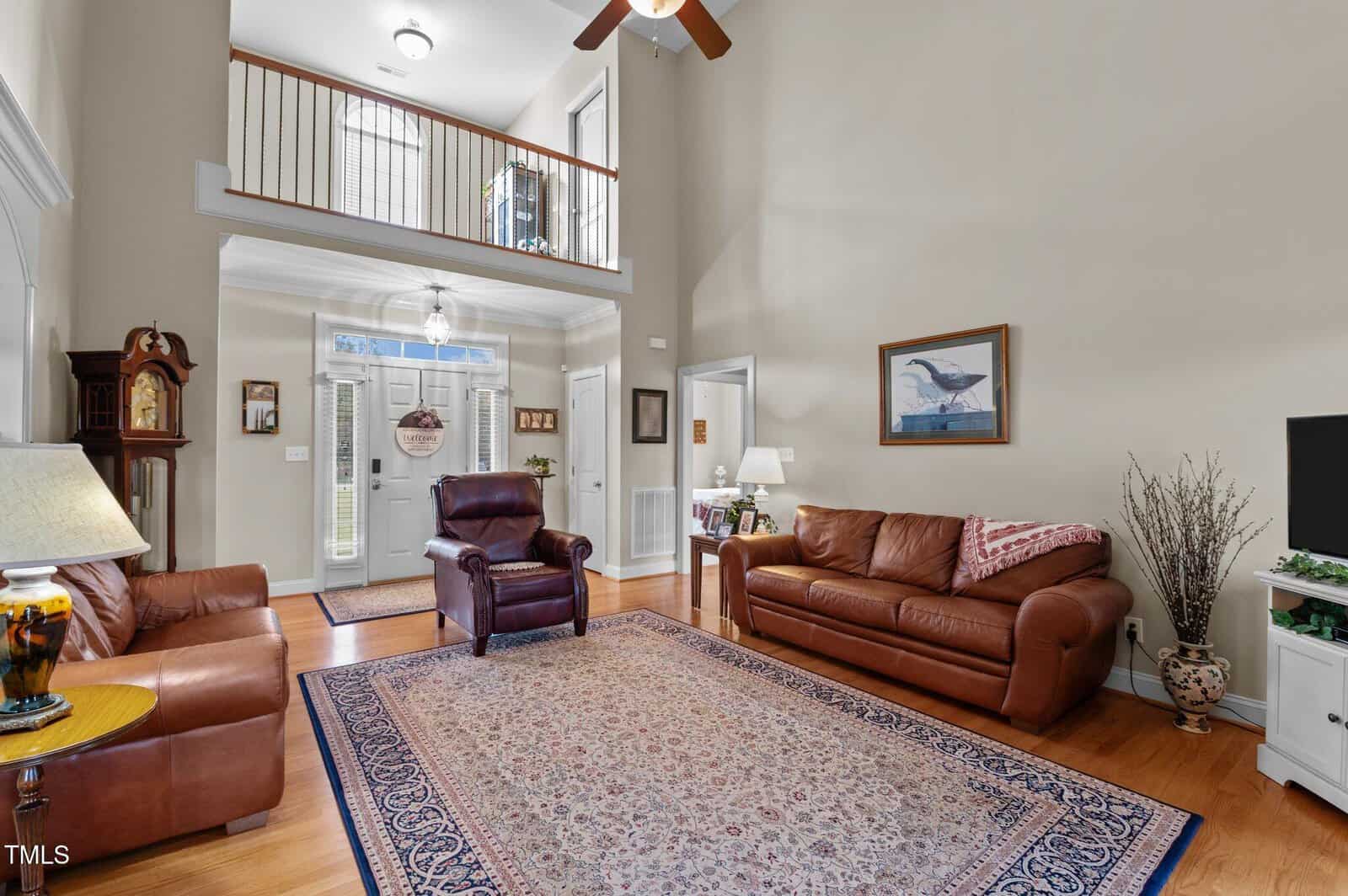 A living room with high ceilings and hardwood floors features leather couches, a plush chair, a decorative rug, a grandfather clock, and a TV. A framed picture hangs on the wall, and the entryway with a window above the door is visible.