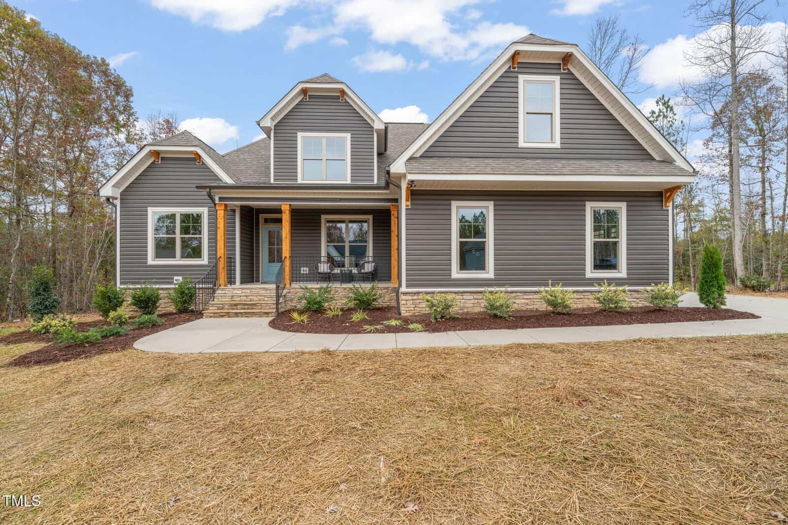 A modern two-story house with gray siding, white trim, and a covered front porch. The yard features new landscaping with small shrubs and mulch, and the driveway curves in front of the home.