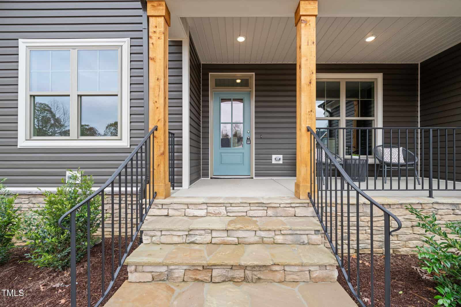 Front porch of a house with stone steps, a light blue door, dark gray siding, wooden columns, black railings, and a small seating area to the right. Bushes line the walkway and porch area.