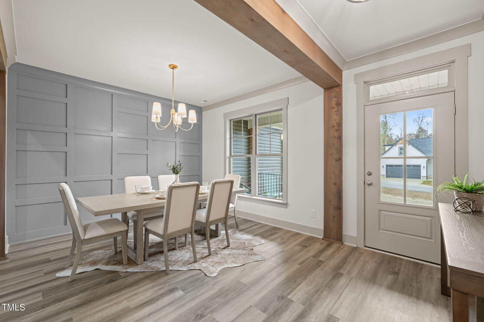 A dining room with a light wood table, six upholstered chairs, a modern chandelier, and a large window. The space features light gray paneled walls, wood floors, and a glass door leading outside.