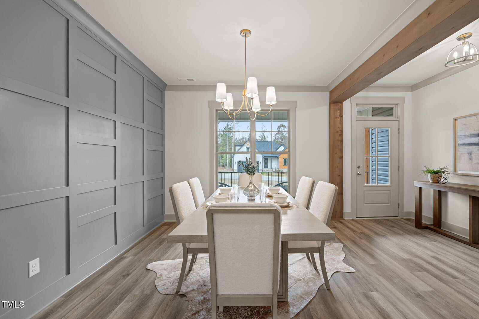 Modern dining room with a rectangular table, six upholstered chairs, geometric accent wall, chandelier, large window, and wood flooring. A cowhide rug is under the table, and a wood beam frames the space near the entry.