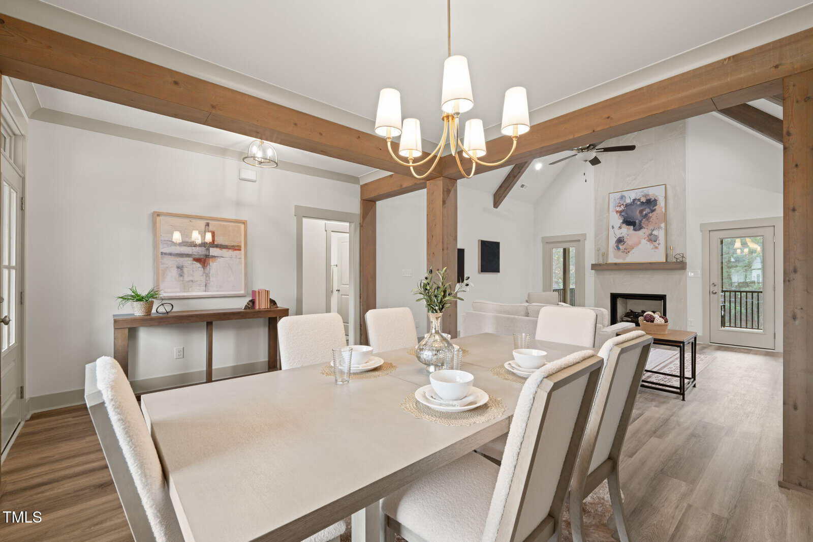 A bright dining room with a light wood table set for four, upholstered chairs, a modern chandelier, exposed wood beams, and an open view into a living area with a fireplace and wall art.