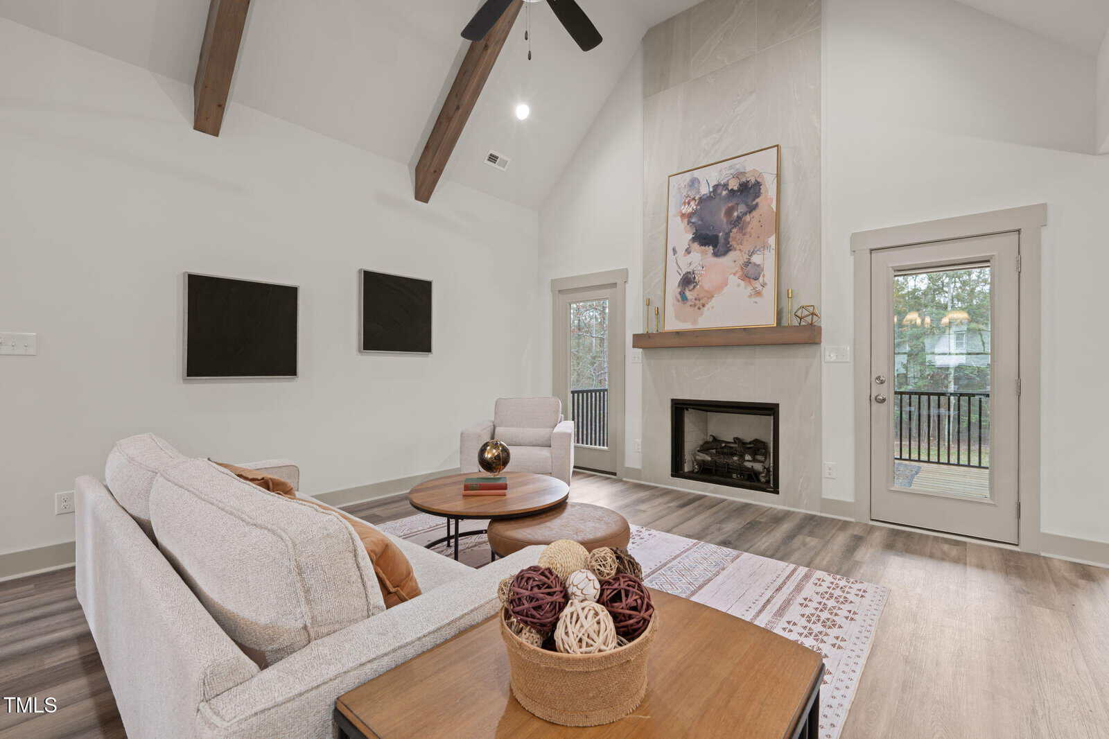 A modern living room with light furniture, a fireplace, abstract art above the mantel, and a glass door leading to a balcony. Exposed wooden beams accent the high ceiling, and natural light fills the space.