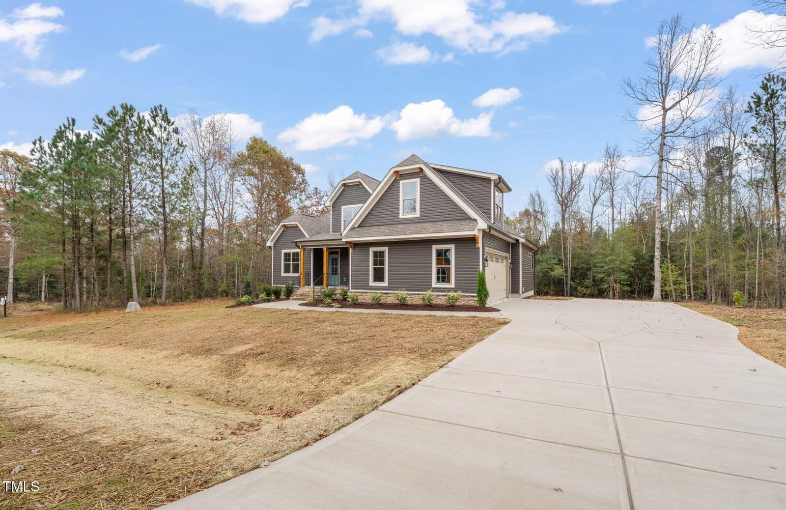 A two-story house with gray siding, white trim, and a covered front porch, sits on a large lot with a wide driveway, minimal landscaping, and surrounded by trees under a partly cloudy sky.