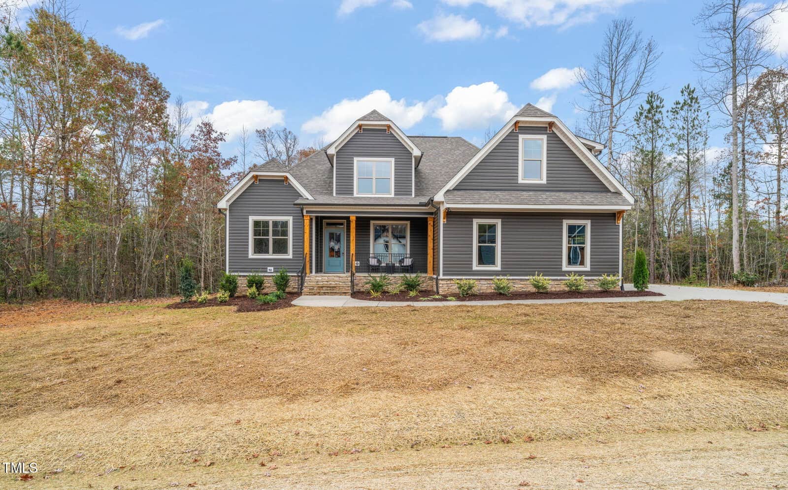 A modern two-story house with gray siding, white trim, a covered front porch, stone accents, and a gabled roof, set on a freshly landscaped lot with surrounding trees under a partly cloudy sky.