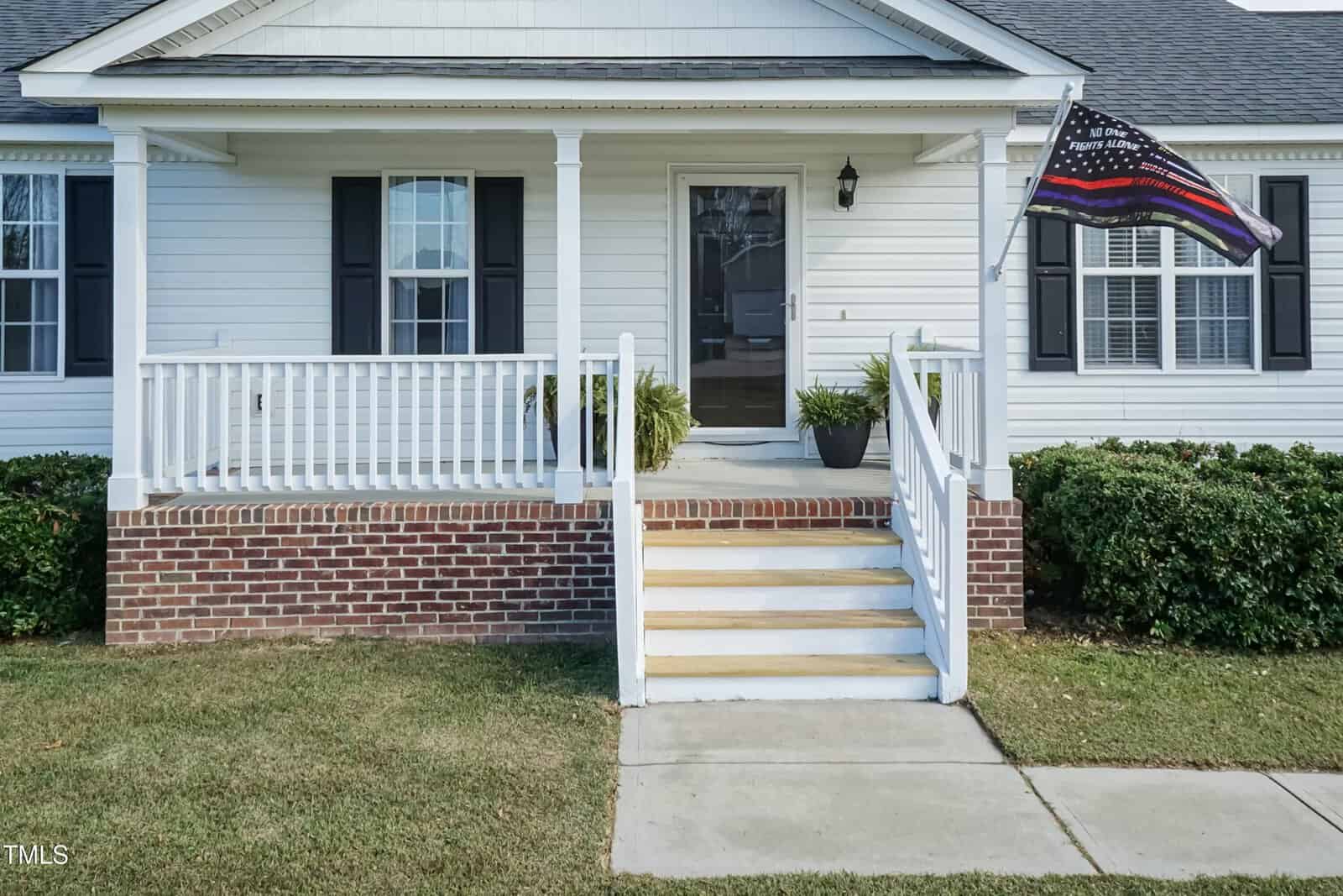 A white house with a small front porch, black shutters, and a multicolored flag. Brick steps lead up to the porch, with potted plants on either side of the entrance, surrounded by green shrubs and grass.