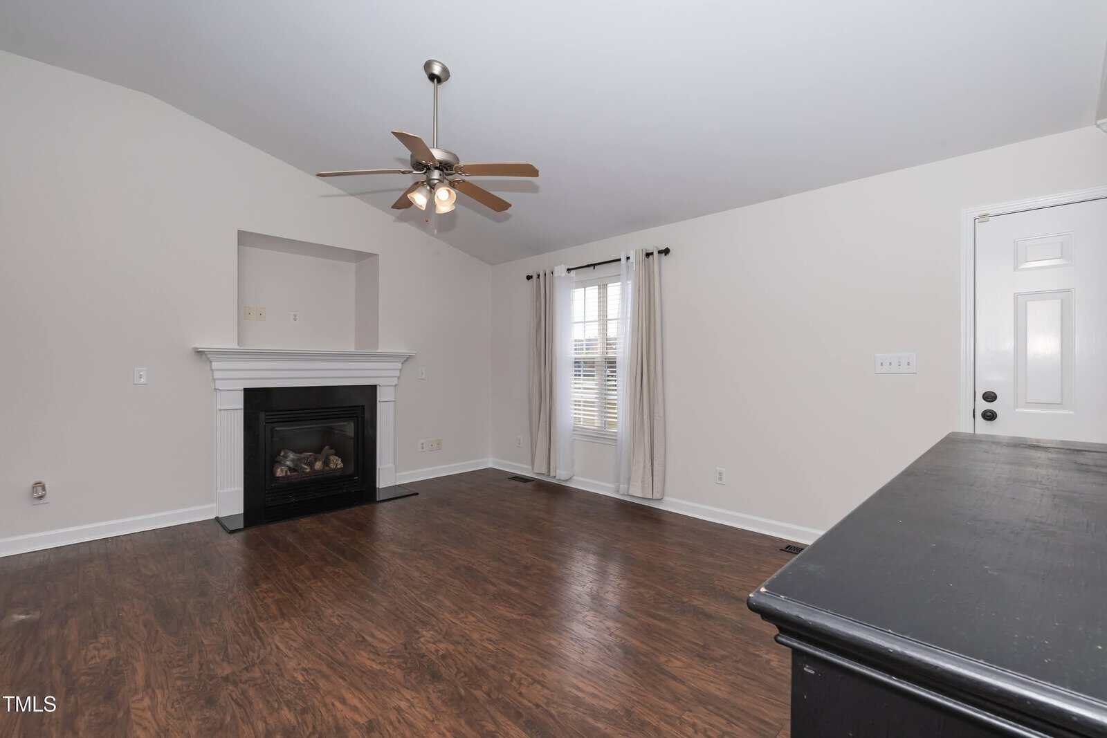 A bright, empty living room with hardwood floors, a ceiling fan with lights, a white fireplace, a window with sheer curtains, and a white door. The walls are painted light gray.