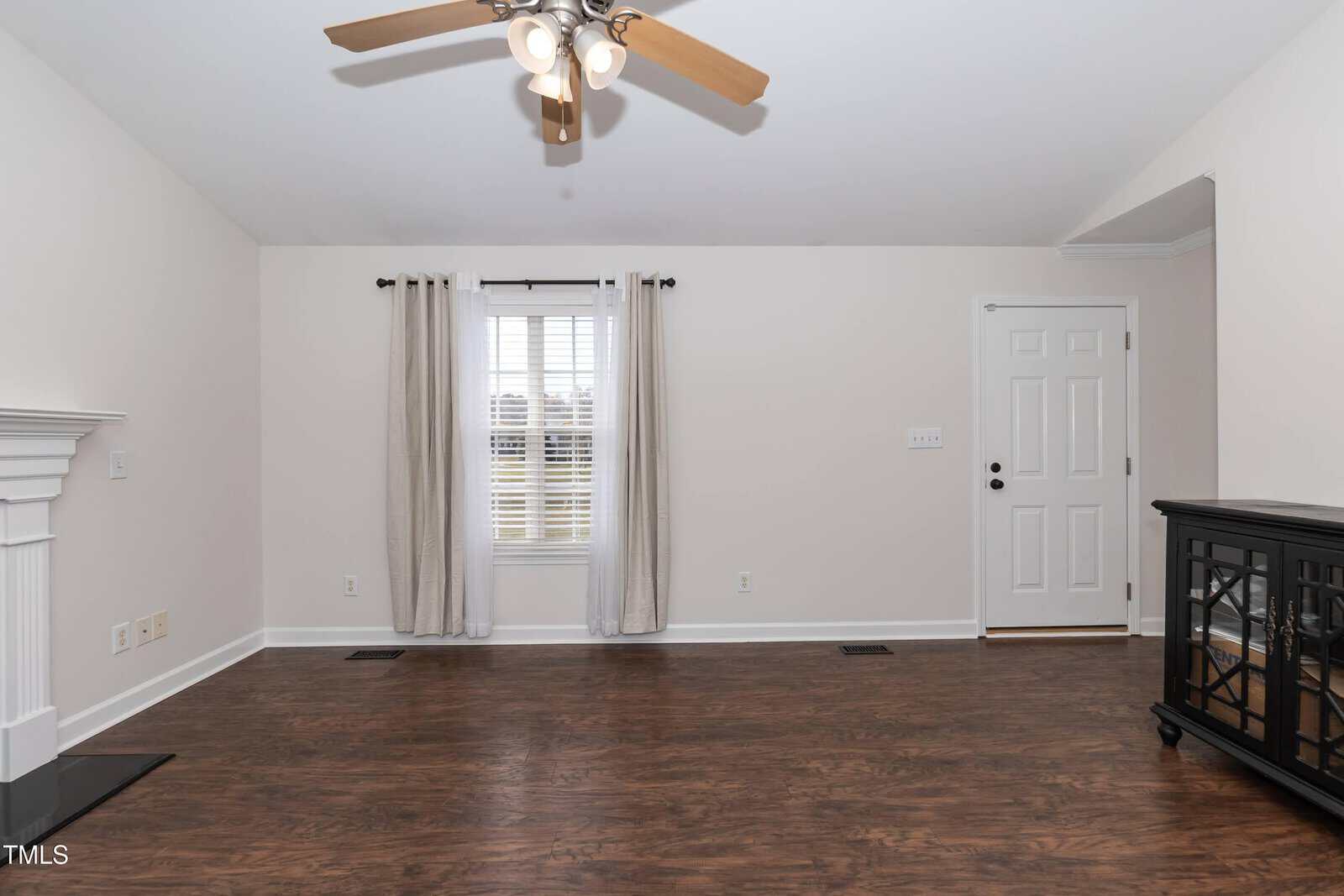 A bright, empty living room with wood flooring, white walls, a ceiling fan with lights, a window with white curtains, a white front door, and a black side cabinet.