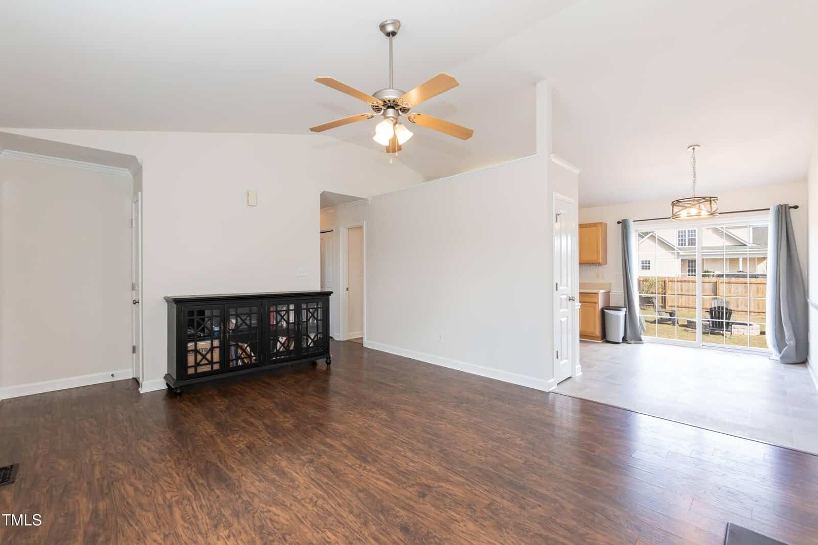 A bright living and dining area with wood flooring, white walls, a ceiling fan, black sideboard, and an open view into the kitchen and backyard through sliding glass doors.