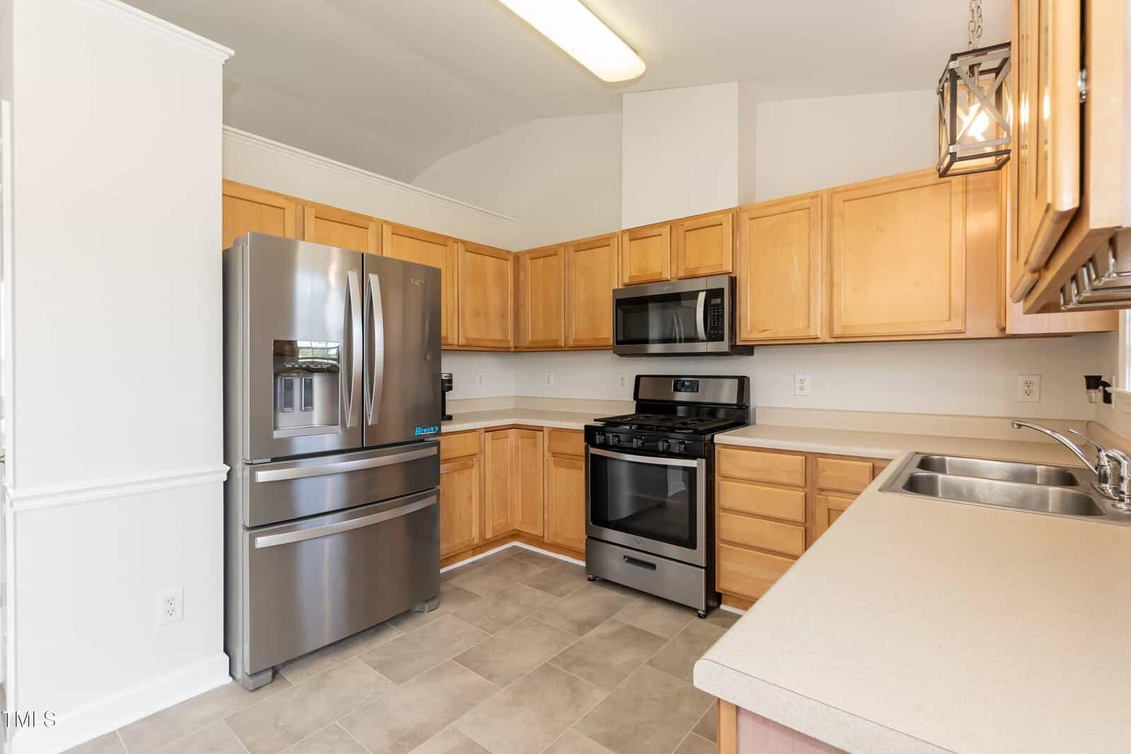 A modern kitchen with light wood cabinets, stainless steel appliances including a refrigerator, oven, and microwave, beige countertops, tiled floor, and bright overhead lighting.