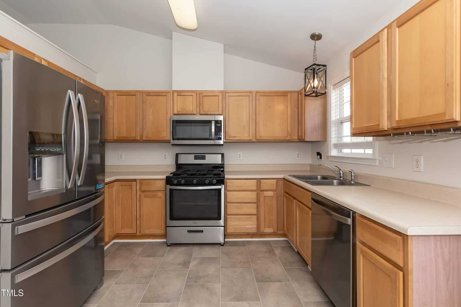 Modern kitchen with stainless steel appliances, including a refrigerator, gas stove, microwave, and dishwasher. Light wood cabinets, beige countertops, tile floor, and a window above the sink.