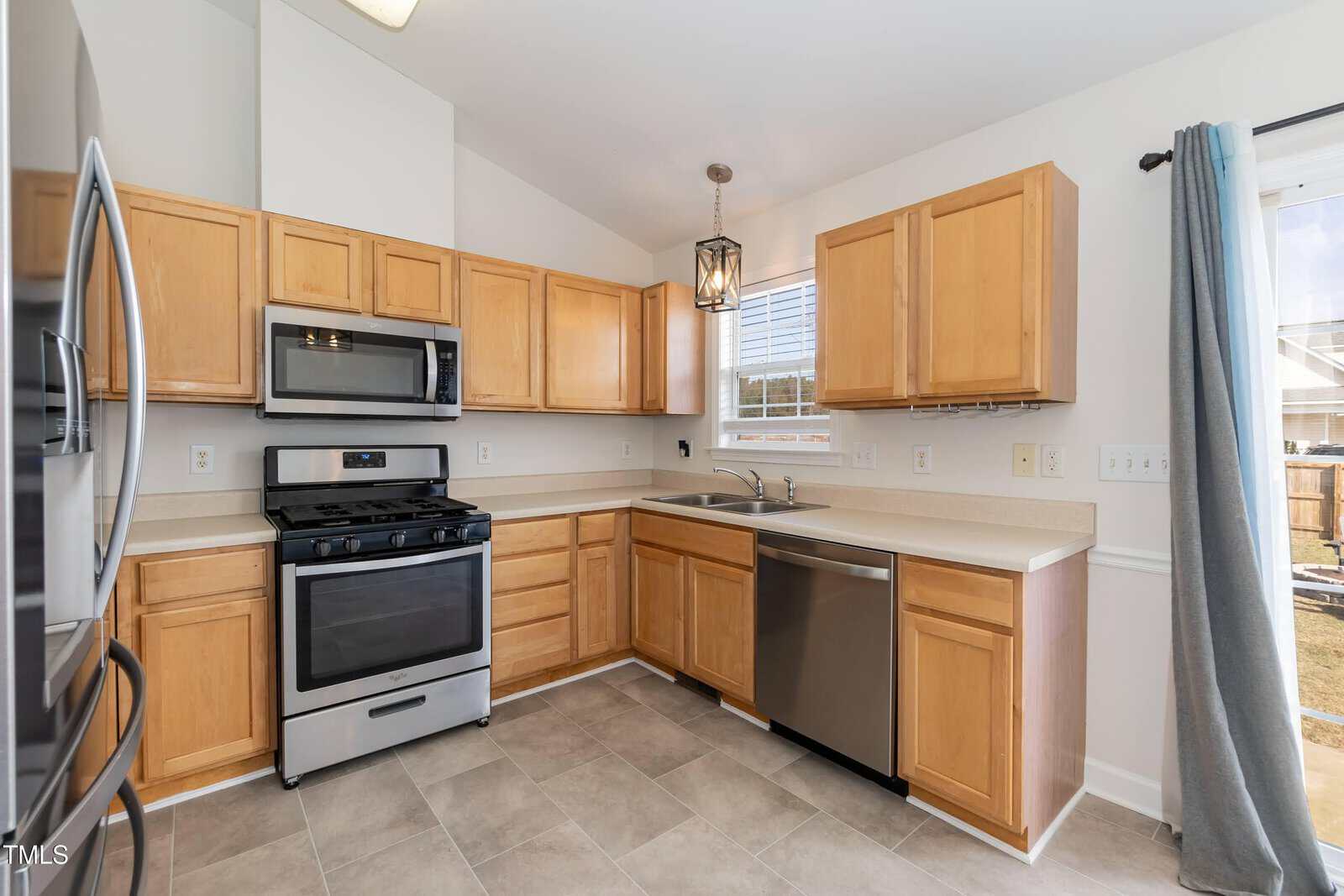 Kitchen with light wood cabinets, stainless steel appliances, beige countertops, dual sink, and tile flooring. There is a window above the sink and a blue curtain covering a glass door to the right.