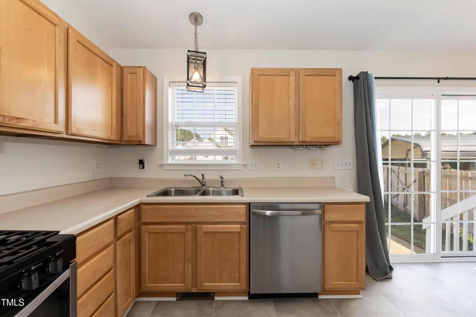 Kitchen with light wood cabinets, beige countertops, stainless steel dishwasher, and stove. Sink is under a window; sliding glass doors with gray curtains lead outside. Walls are white, and floor is tiled.