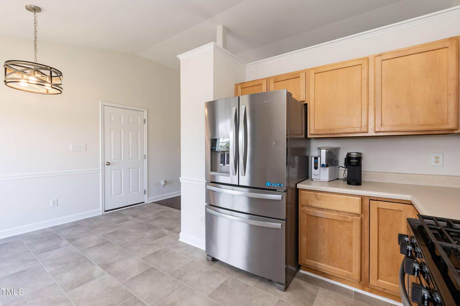 Modern kitchen with stainless steel refrigerator, wooden cabinets, coffee maker, and beige tiled floor. A door and a window are visible, with a round light fixture hanging above the open space.